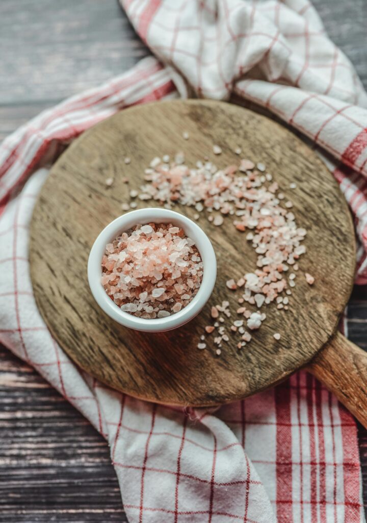 A bowl of pink Himalayan salt on a wooden board surrounded by a checkered cloth, viewed from above.